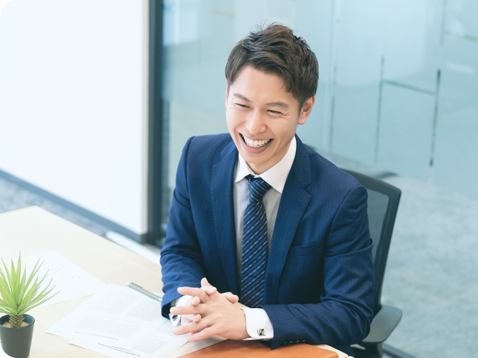 明るいオフィスで、青いスーツを着た笑顔のビジネスマンがデスクに座り、手を組んでいる様子。楽しそうでプロフェッショナルな営業の雰囲気